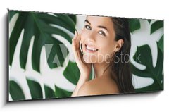 Skleněný obraz 1D panorama - 120 x 50 cm F_AB217474275 - Portrait of young and beautiful woman with perfect smooth skin in tropical leaves - Portrét mladé a krásné ženy s dokonalou hladkou pokožkou v tropických listech